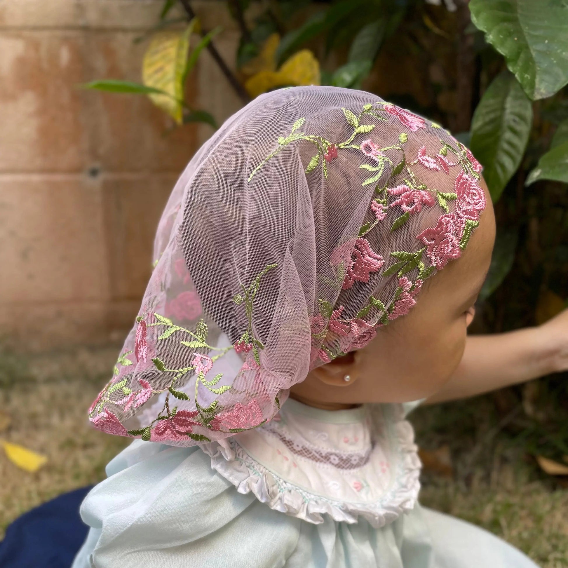 Baby wearing a traditional pink embroidered chapel veil for Christian ceremony.