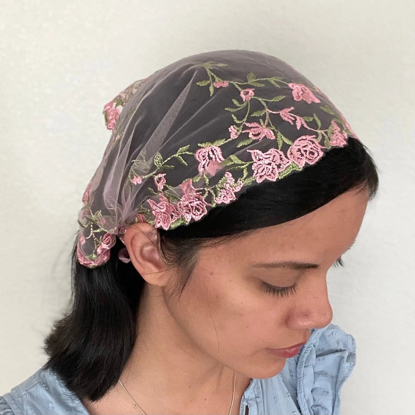 Girl wearing a pink lace chapel veil with rose embroidery. The small Traditional Catholic Christian Mass mantilla has ties