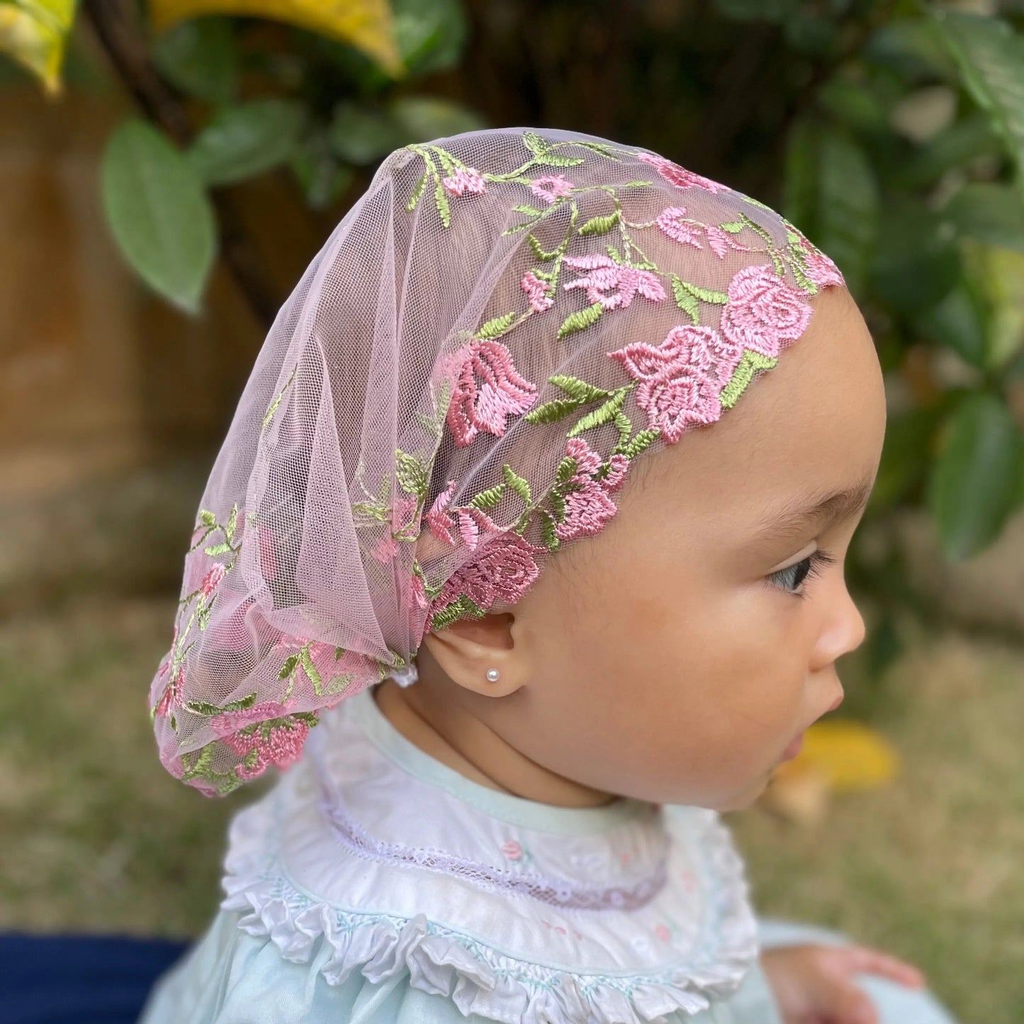 Infant wearing pink embroidered lace veil, a traditional Catholic chapel veil.