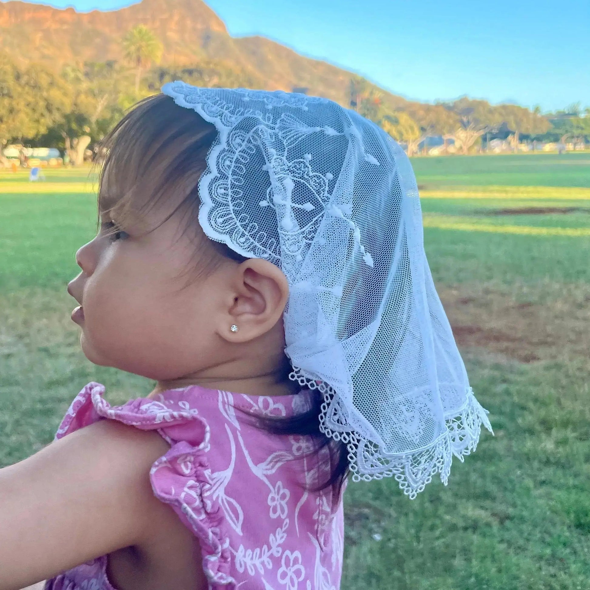 A baby wearing a white lace baptism veil with embroidered crosses, featuring a D-shaped cut.
